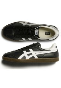 Černé kožené tenisky s bílými detaily, které mají vrstevnatý design bočního pruhu, černé tkaničky a gumovou podrážku. Klasický atletický styl.