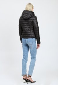 Sort puffervinterjakke med hætte, quiltet design og blank tekstur, parret med blå jeans og sorte hælede sandaler.