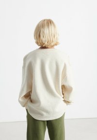 Krämfärgad fleece-tröja med avslappnad passform, rundad nederkant och droppe axlar. I kombination med gröna byxor, sedd bakifrån.