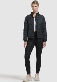 Čierna prešívaná bunda s vysokým golierom a zipovým zapínaním, nosená cez biele crop top a čierne legíny. Neformálna športová obuv.