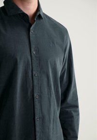 Donkergroene button-up shirt van zachte stof met een kraag, voorzien van een tonale geborduurde logo en bruine knopen aan de voorkant.