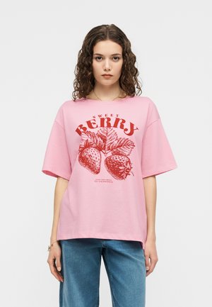Junge Frau mit lockigem Haar, die ein pinkfarbenes, übergroßes T-Shirt mit rotem Erdbeermotiv und blaue Jeans trägt, vor einem schlichten Hintergrund steht.