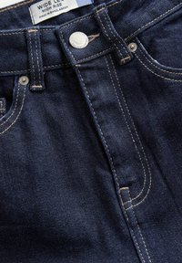 Jean denim bleu foncé, taille haute, jambe large, bouton en métal et coutures contrastées, étiquette indiquant fabriqué au Bangladesh.