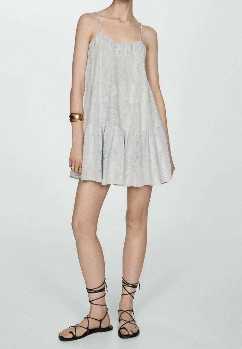 Personne portant une robe ample sans manches de couleur gris clair avec des bretelles fines, associée à des sandales plates noires à lacets et des bracelets dorés au poignet gauche.