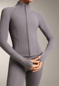 Šedá atletická bunda na zips, vyrobená z hladkej tkaniny, s vysokým golierom, priliehavým strihom a otvormi na palce na manžetách rukávov pre pohodlie.