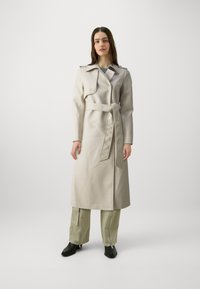 Beige trenchcoat met een ceintuur in de taille, dubbelgeknoopte knopen en schouderepauletten. Combinatie met olijfgroene cargobroeken en zwarte enkellaarsjes.