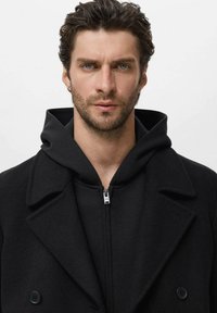 Cappotto peacoat nero sopra una felpa nera con zip. Il peacoat presenta grandi bottoni e un colletto a risvolto; trame distinte del tessuto sono visibili.