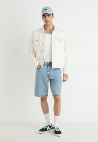 Veste en denim blanc sur un débardeur blanc, associée à un short en denim bleu clair. Les accessoires comprennent une casquette bleu clair et des baskets navy.