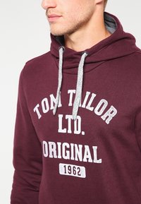 Bordeauxrode hoodie met grijze treklussen, voorzien van een gedrukt logotekst "TOM TAILOR LTD. ORIGINAL 1962" in wit. Casual ontwerp, zachte textuur.