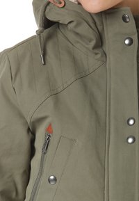 Olijfgroene jas met een hoge kraag, voorzien van een afneembare capuchon, ritszak en decoratief oranje driehoeklabel. Gladde textuur en drukknoopsluitingen.