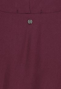 Nahaufnahme eines lila Stoffes, auf dem ein kleiner runder Metallknopf mit Näharbeit und eingraviertem Markennamen zu sehen ist.