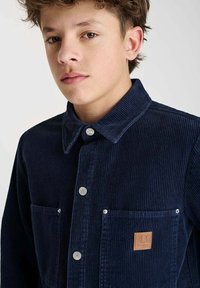 Marineblauwe ribfluwelen blouse met zilveren knopen, twee voorzakken en een leren logo-patch. Voorzien van een klassieke kraag en ribbelstructuur.