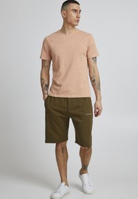 Tricou scurt de culoare roz deschis, cu mânecă scurtă, asortat cu pantaloni scurți de culoare verde oliv, dotat cu buzunare laterale, textură netedă și adidași albi.