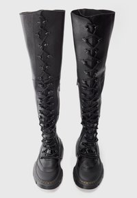 Black leather thigh-high lace-up boots featuring metal eyelets, a rounded toe, textured panels, and a chunky rubber sole.