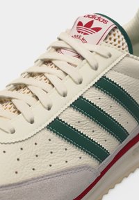 Baskets Adidas blanc cassé avec des bandes vertes, des lacets beige, un logo rouge sur la languette, des côtés perforés et un renfort de gomme suédé à l'avant.