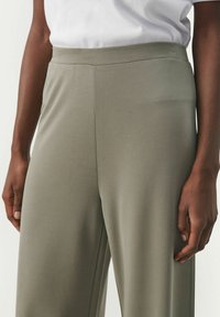 Pantalons larges vert olive en tissu lisse, avec une taille haute et une silhouette ajustée, présentés avec un t-shirt blanc.