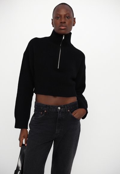 Black cropped sweater with high collar and silver zipper, paired with dark denim jeans featuring front pockets and copper buttons.