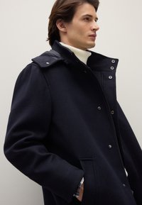 Cappotto in misto lana blu navy con collo alto, chiusura con zip e bottoni a pressione. Presenta tasche laterali e polsini di una camicia a quadri visibili sotto.