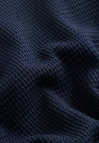 Tamsiai mėlyna megzto audinio medžiaga, išsiskirianti tekstūruotu, tinklelio tipo raštu su skirtingais pakilimais, demonstruojanti minkštą ir šiek tiek elastingą paviršių.