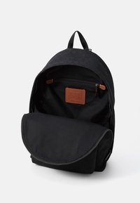 Sac à dos en tissu noir avec un dessus texturé, une fermeture éclair et un rabat inférieur lisse. Comprend un écusson de marque en cuir marron à l'intérieur.