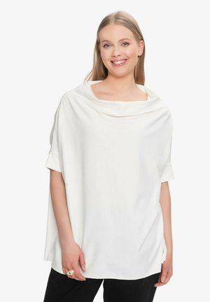 Uśmiechnięta kobieta w luźnej, białej bluzce z krótkim rękawem i szerokim dekoltem typu komin, w połączeniu z czarnymi spodniami, stojąca na białym tle.