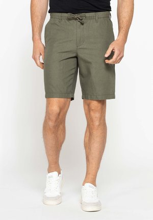 Uomo che indossa pantaloncini verde oliva al ginocchio con coulisse, maglietta nera e sneakers bianche, in piedi su uno sfondo semplice.