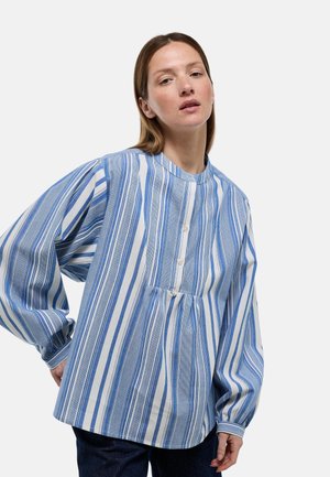 Femme portant un chemisier ample bleu et blanc à rayures verticales avec de longues manches bouffantes et un col rond, debout devant un fond uni.