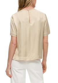 Blouse beige à manches courtes avec un léger éclat, dotée d'une ouverture en trou de serrure dans le dos, et d'une coupe droite à l'ourlet. Portée avec un pantalon blanc.