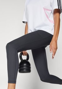 Melnas ribotas legingi ar augstu jostasvietu un baltu brīvi plūstošu kreklu ar rozā akcentiem. Vienā rokā tur melnu kettlebellu.
