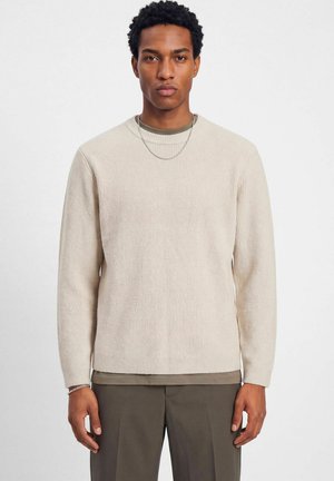 Un homme porte un pull côtelé couleur crème par-dessus une chemise taupe, avec un pantalon foncé et un collier en argent.