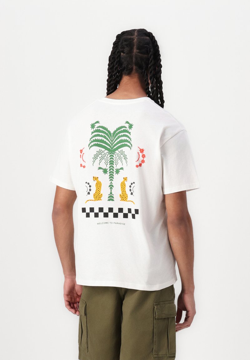 Hvid bomulds t-shirt med et farverigt palmetræ grafik, to leoparder, røde og grønne accenter, og et ternet mønster nederst.