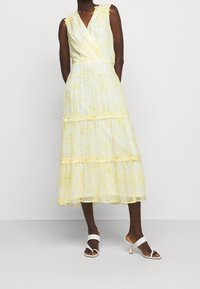Gul klänning i omlottskärning med ett texturerat mönster, volangdetaljer och en flerlagrig kjol. Matchad med vita klacksandaler.