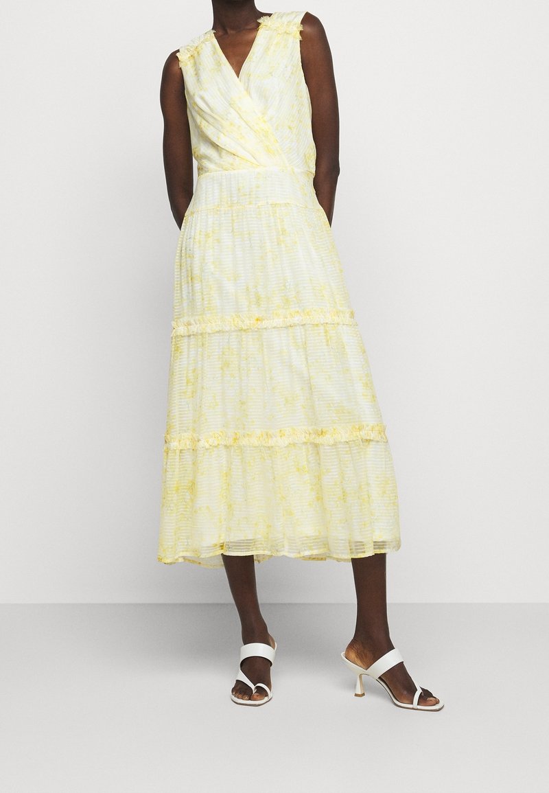 Gul klänning i omlottskärning med ett texturerat mönster, volangdetaljer och en flerlagrig kjol. Matchad med vita klacksandaler.