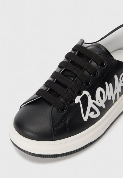 Sneaker en cuir noir avec semelle en caoutchouc blanche, dotée de lacets noirs et d'un logo en style graffiti blanc sur le côté. Texture lisse.