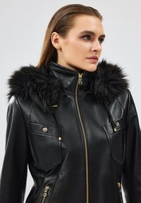 Giacca di pelle nera con cappuccio in pelliccia sintetica, chiusura frontale con zip e accessori in tonalità oro. Include due tasche sul petto e finitura testurizzata.