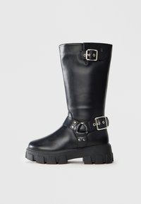 Botine negre din piele până la jumătatea gambei, cu talpă groasă tip lug, două curele cu cataramă argintie — una în partea de sus și una în jurul gleznei.