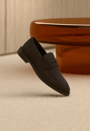 Μαύρο δερμάτινο loafer χωρίς κορδόνια που αιωρείται πάνω από μπεζ χαλί με ένα στρογγυλό τραπεζάκι σε κεχριμπαρένιο καφέ χρώμα στο φόντο.