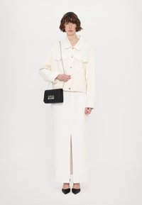 Giacca di denim bianca con bottoni argento, indossata sopra una gonna bianca alla lunghezza del ginocchio con una profonda apertura frontale. Borsa in pelle nera con testo "LOVE MOSCHINO".