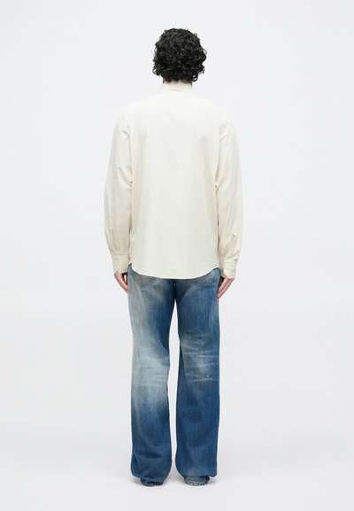 Gaiši bēša krekla ar garām piedurknēm ar pogām uz aprocēm, valkājams ar brīvā piegriezuma zilām džinsa biksēm ar izbalējušu mazgāšanas efektu un platu kāju dizainu.