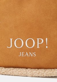 Hnědý semišový pohlavník se bílým vyšitým textem "JOOP! JEANS" a béžovým texturovaným fleecovým lemem podél spodního okraje.