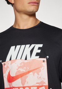 Svart t-skjorte med stor hvit "NIKE" trykt på forsiden, med et rødt mønster under og detaljer i metall.