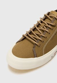 Baskets en toile marron avec bout rond, semelle en caoutchouc crème, lacets marron et couture contrastante. Présente un logo sur la languette et sur le côté.