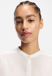 Weiße Hemdbluse mit Stehkragen aus leichtem Stoff, mit leicht transparentem Charakter und dezenten Nähdetails.