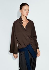 Bruine blouse met kraag, losse pasvorm en lange mouwen, gecombineerd met een donkerblauwe tailleband, voor een neutrale achtergrond.