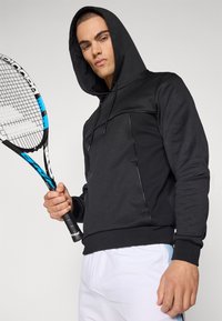 Schwarzer Kapuzensweater mit strukturierten Einsätzen, slim fit und elastischen Bündchen, gehalten von einem Modell, das einen Tennisschläger greift.