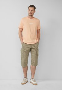 T-shirt crew en pêche clair avec logo sur le devant, associé à un short cargo vert olive avec poches latérales et coupe décontractée. Baskets crème.