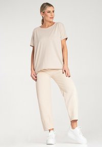 Beżowa bluzka z krótkim rękawem o luźnym kroju, w połączeniu z dopasowanymi spodniami i białymi trampkami. Gładki, miękki materiał z delikatnym połyskiem.