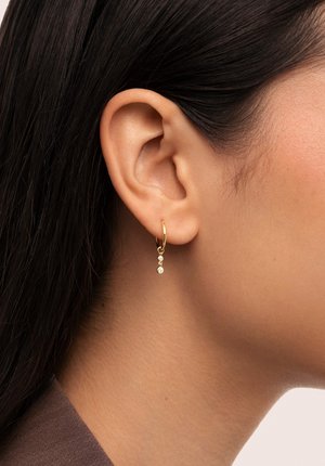Goldene Creolen mit drei kleinen, befestigten klaren Kristallen, die am Ohr eines Modells präsentiert werden. Glatte Textur, kreisförmige Form, dezenter Glanz.
