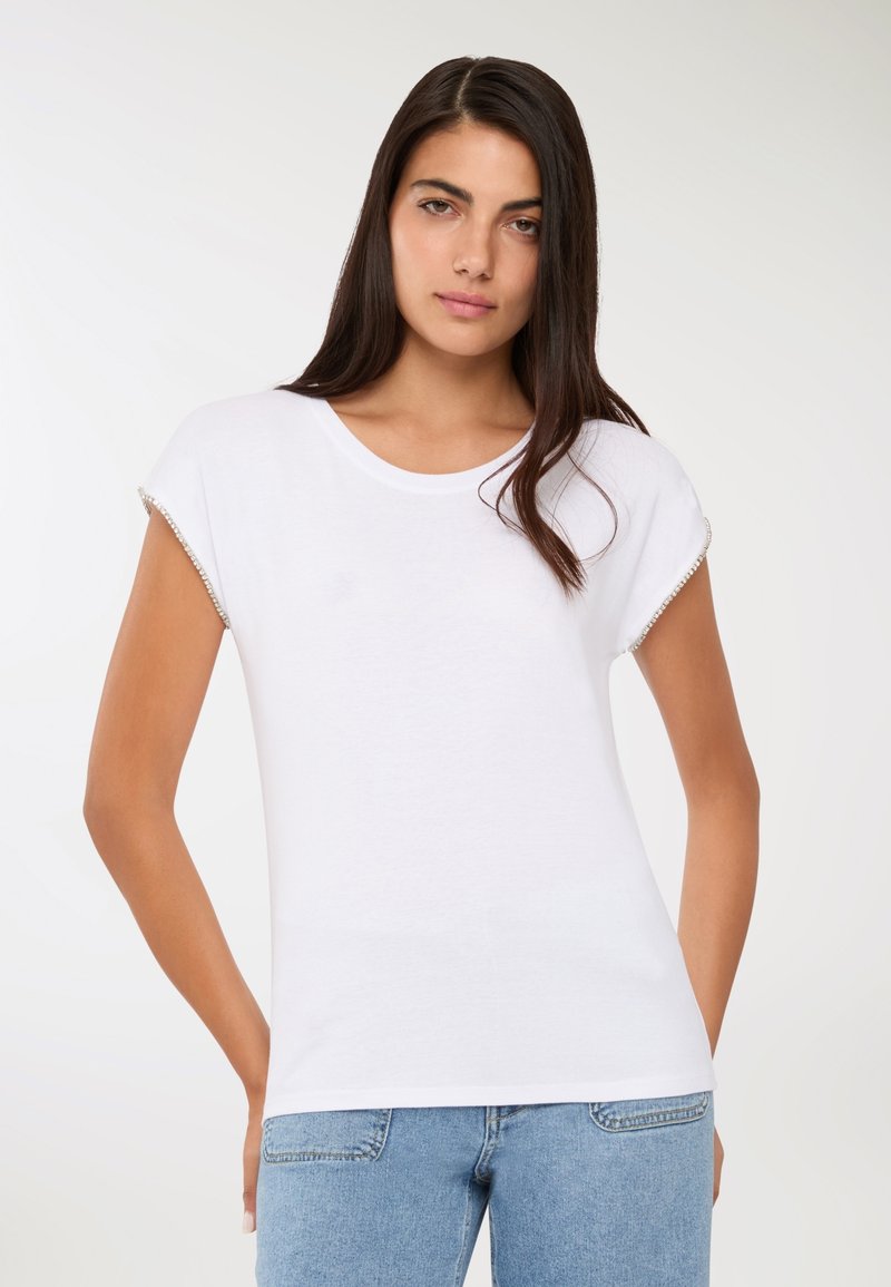 Kobieta z długimi ciemnymi włosami, nosząca biały krótki rękaw top z ozdobnymi krawędziami rękawów i jasnoniebieskie dżinsy, stojąca na jednolitym tle.