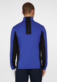 Blå och svart långärmad skjorta med hög krage, som har en vertikal svart varumärkesetikett på baksidan och slät, texturerad tyg.